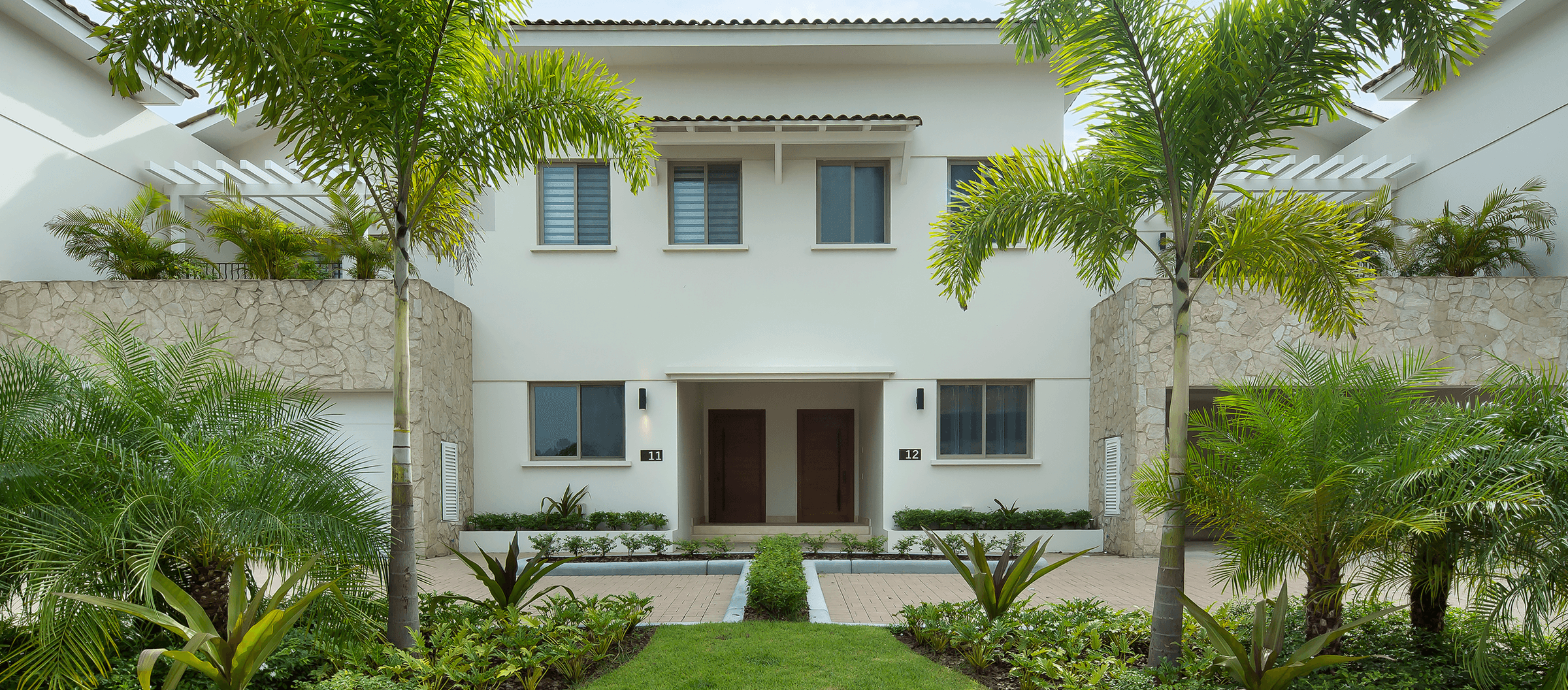 Front of two paired two-storey homes with stone walls and palm-lined entrance garden at The Crescent, Santa Maria