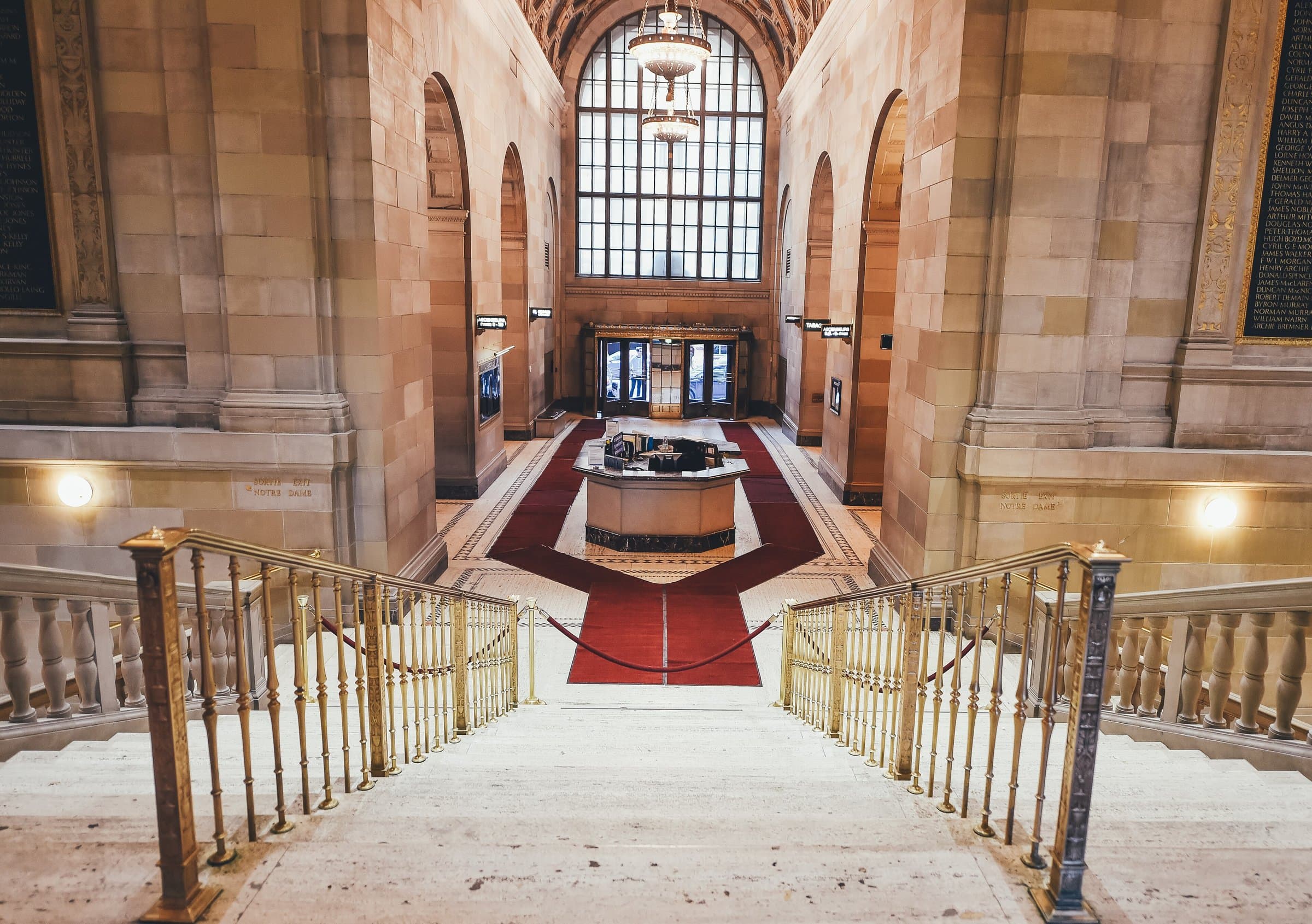 Reception desk at the center of a grand stone entrance hall with red carpet and brass railings