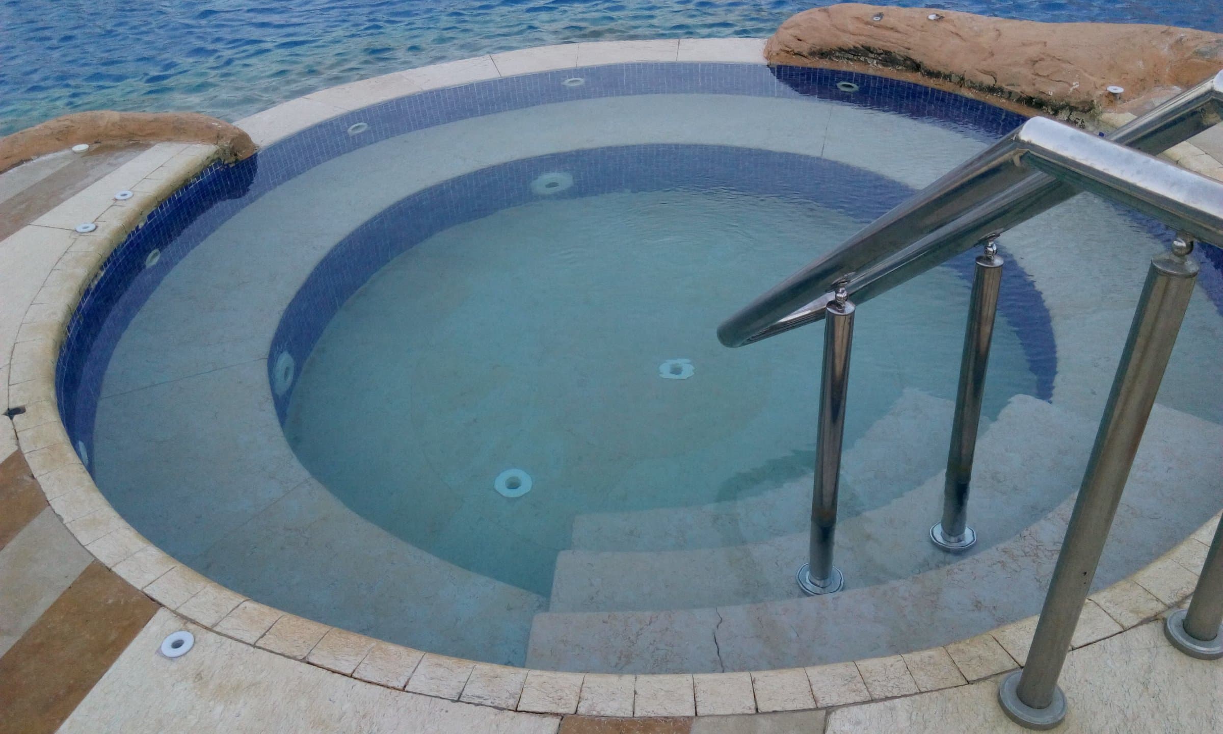 Round outdoor jacuzzi with tiled steps and a steel handrail beside the sea