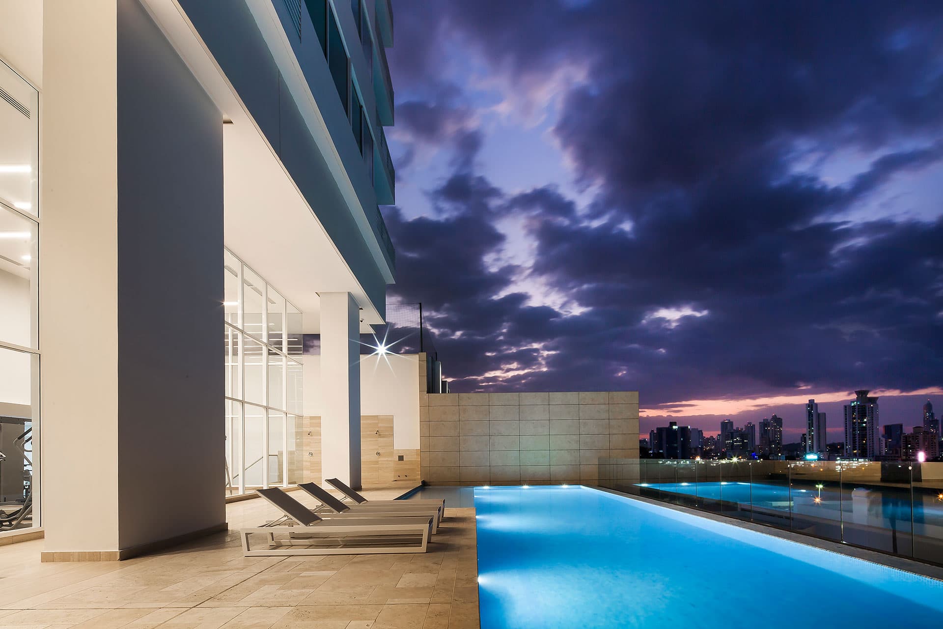 Lit swimming pool with loungers beside the tower at dusk at The Towers Residences