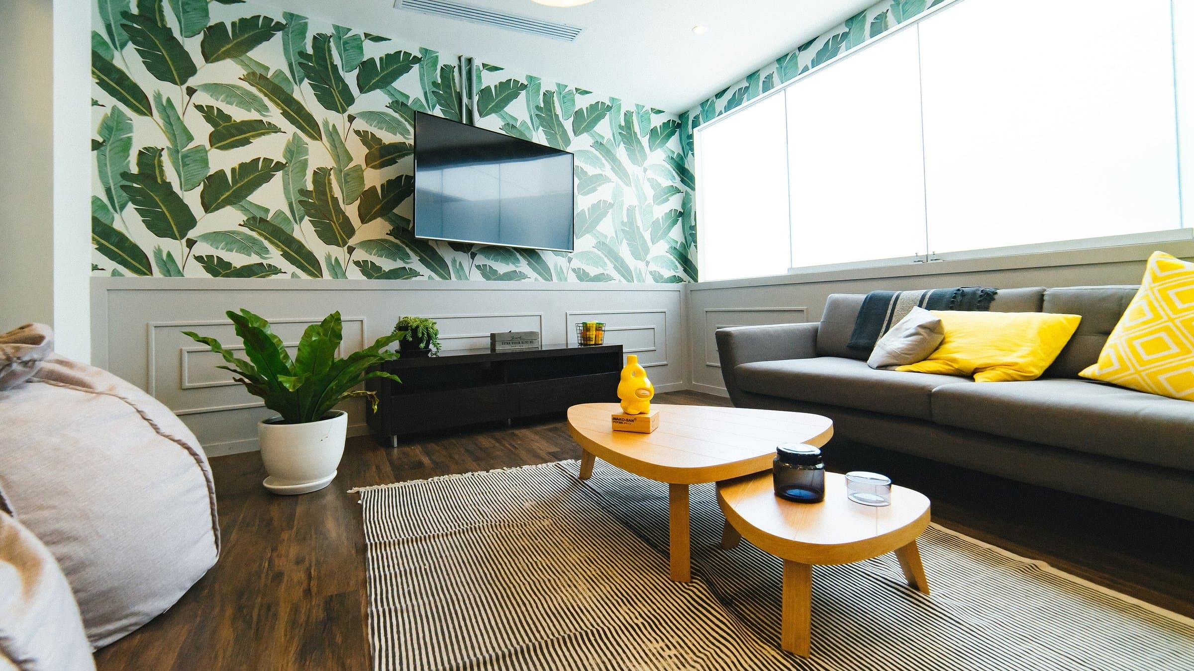 Coworking lounge with a gray sofa, yellow cushions, wooden coffee tables and tropical leaf wallpaper