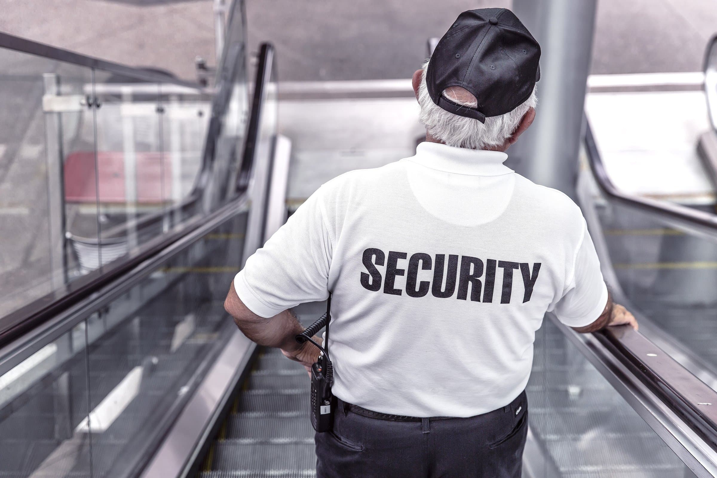 Security guard seen from behind on an escalator, wearing a white uniform shirt and carrying a radio