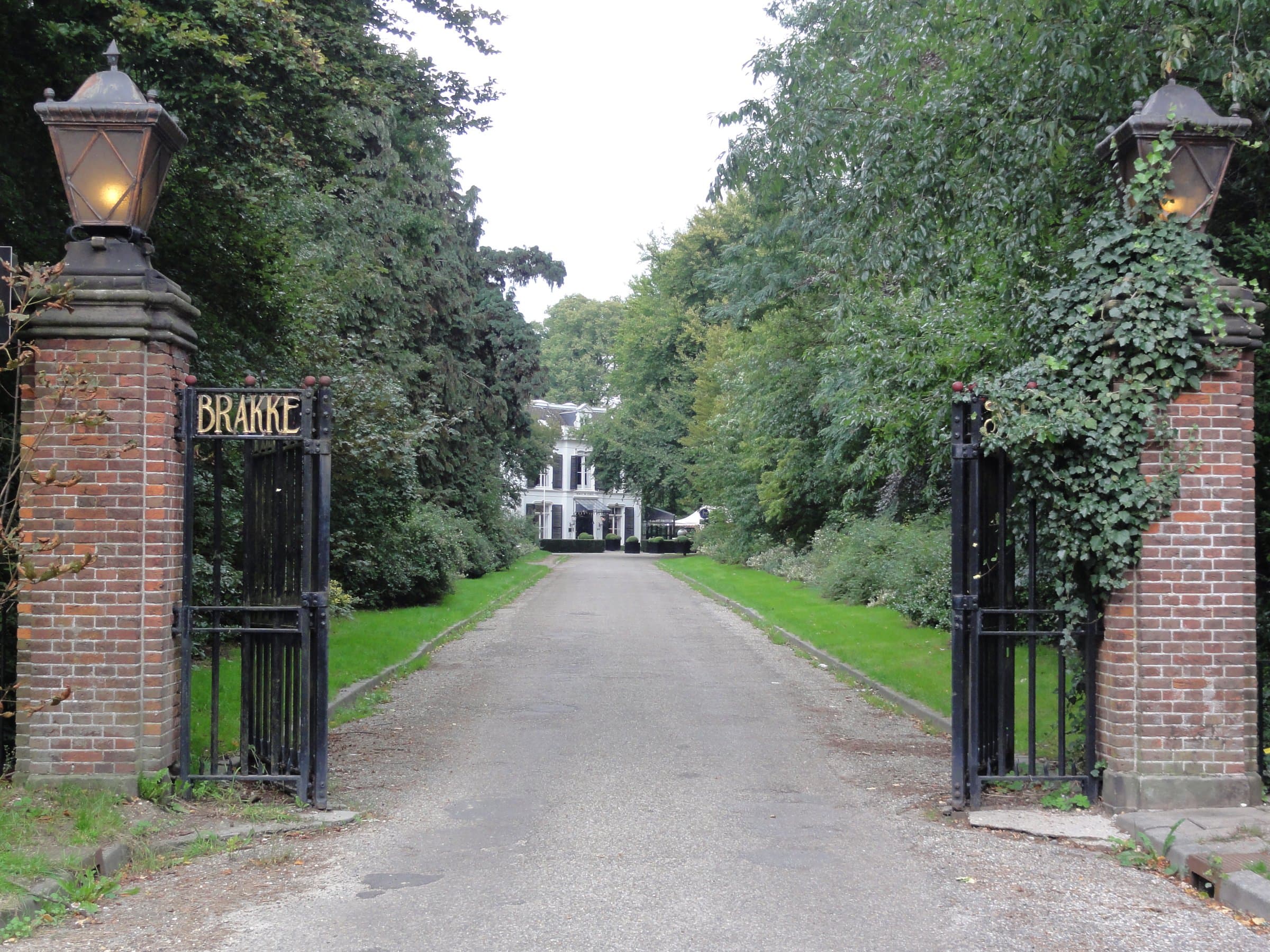 Open wrought-iron gates between brick pillars on a tree-lined drive leading to a white villa