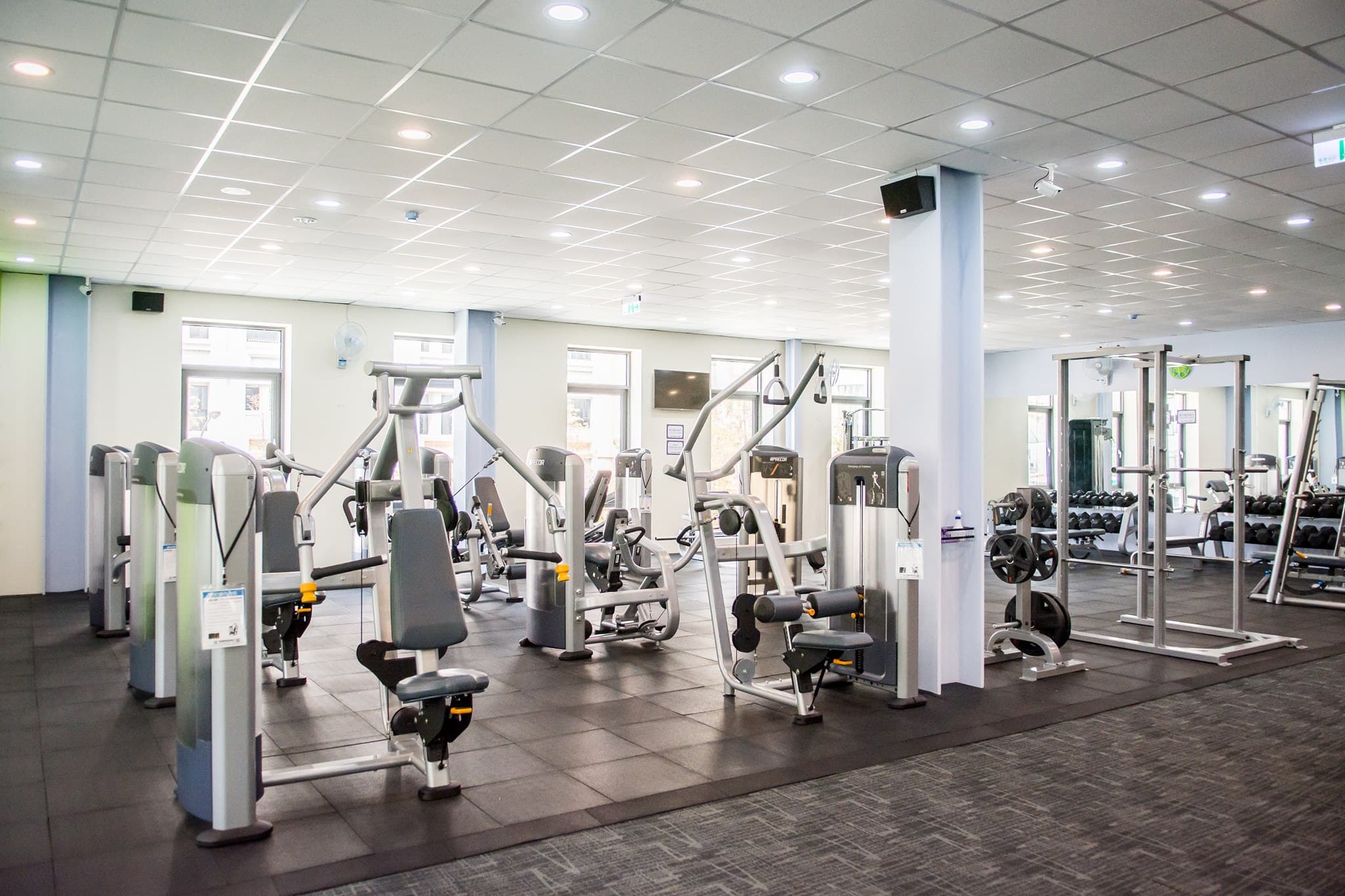 Bright gym with strength machines, a squat rack and dumbbell racks under recessed ceiling lights