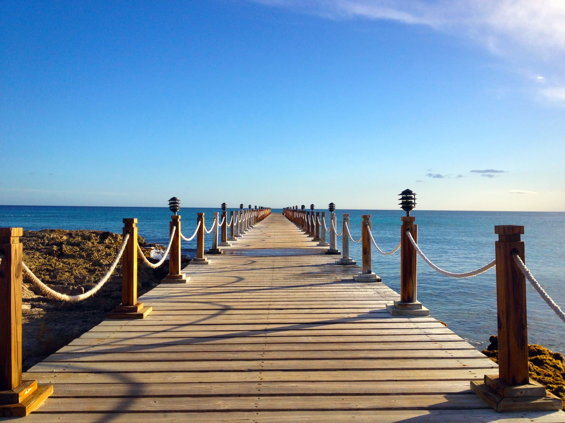 Wooden boardwalk with rope railings leading out over calm blue sea under a clear sky