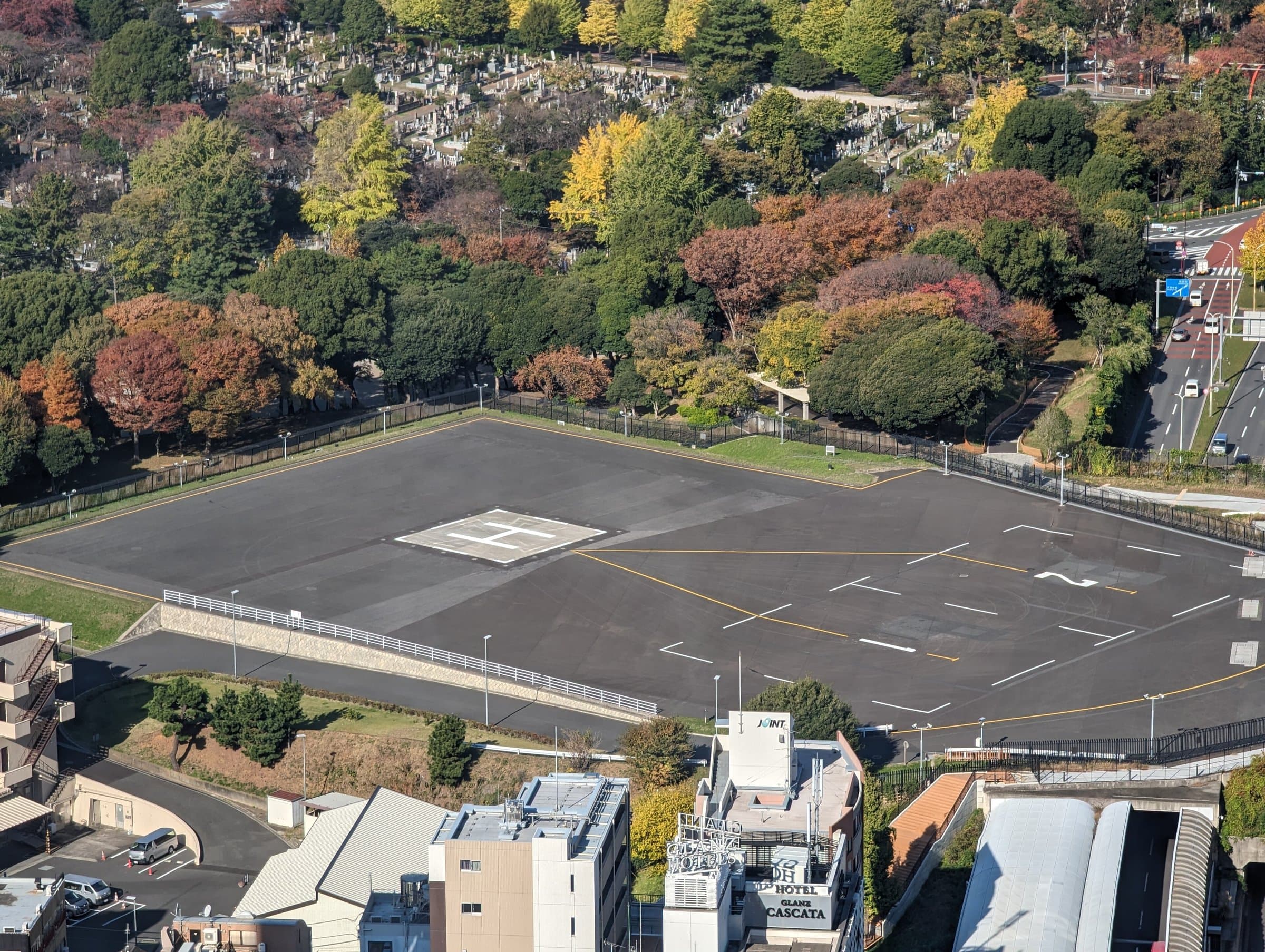 Aerial view of a ground-level helipad with a painted H marking, bordered by a fence and dense trees