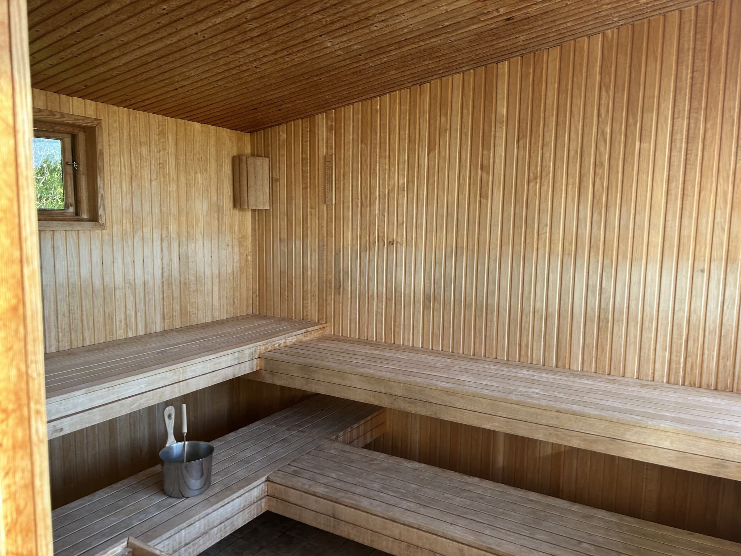 Wood-panelled sauna interior with two tiers of benches, a small window and a bucket with ladle