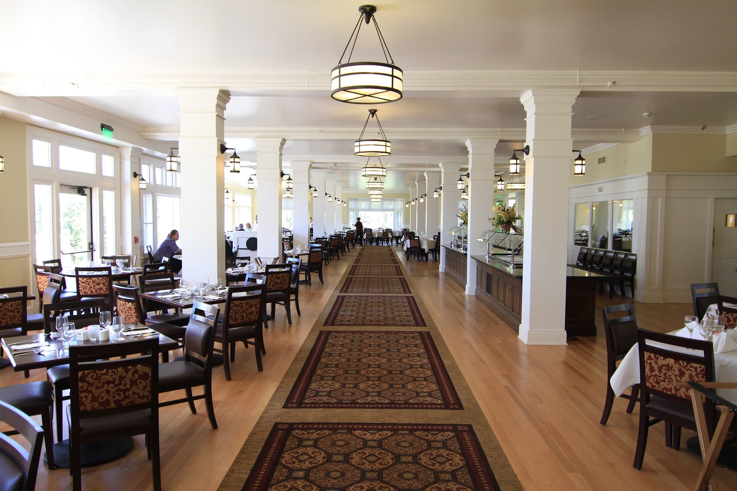 Hotel dining room with white columns, pendant lamps and set tables on either side of a patterned runner