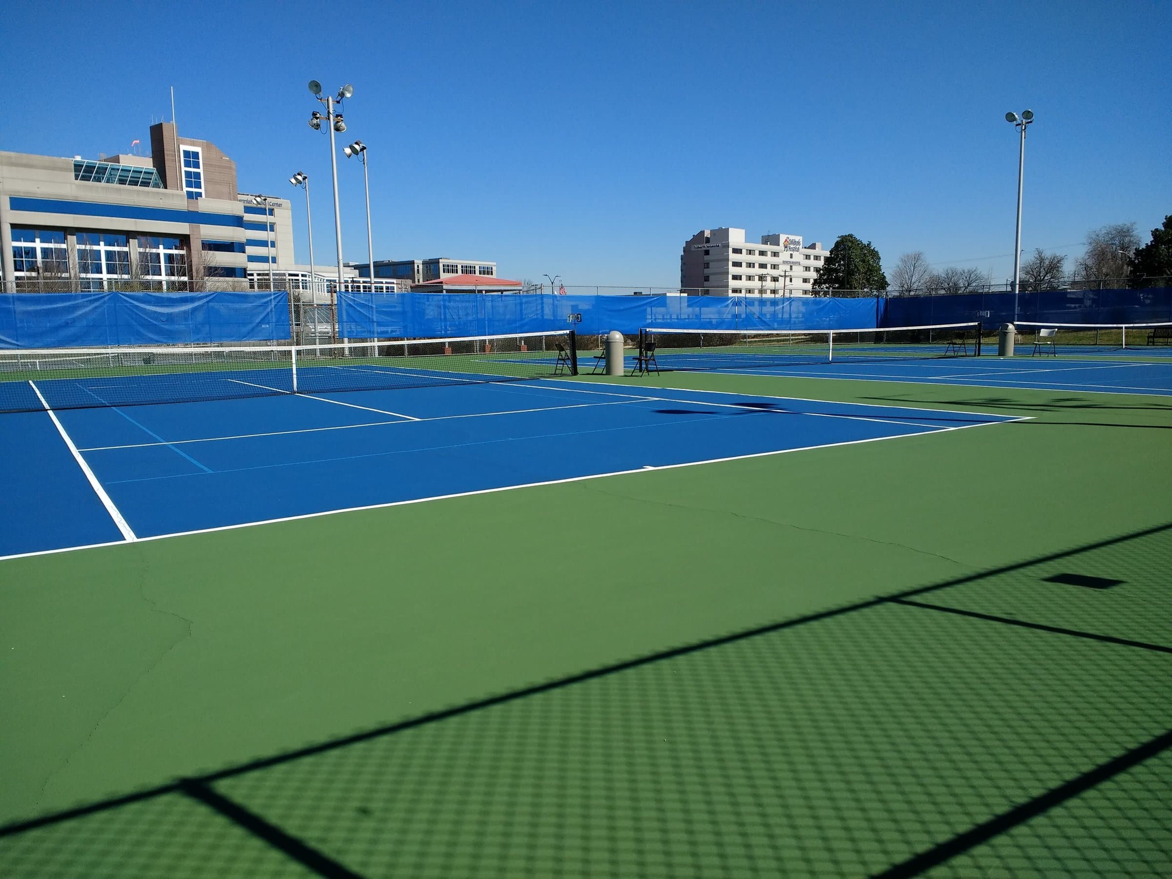 Blue hard tennis courts with nets and green surrounds under a clear blue sky