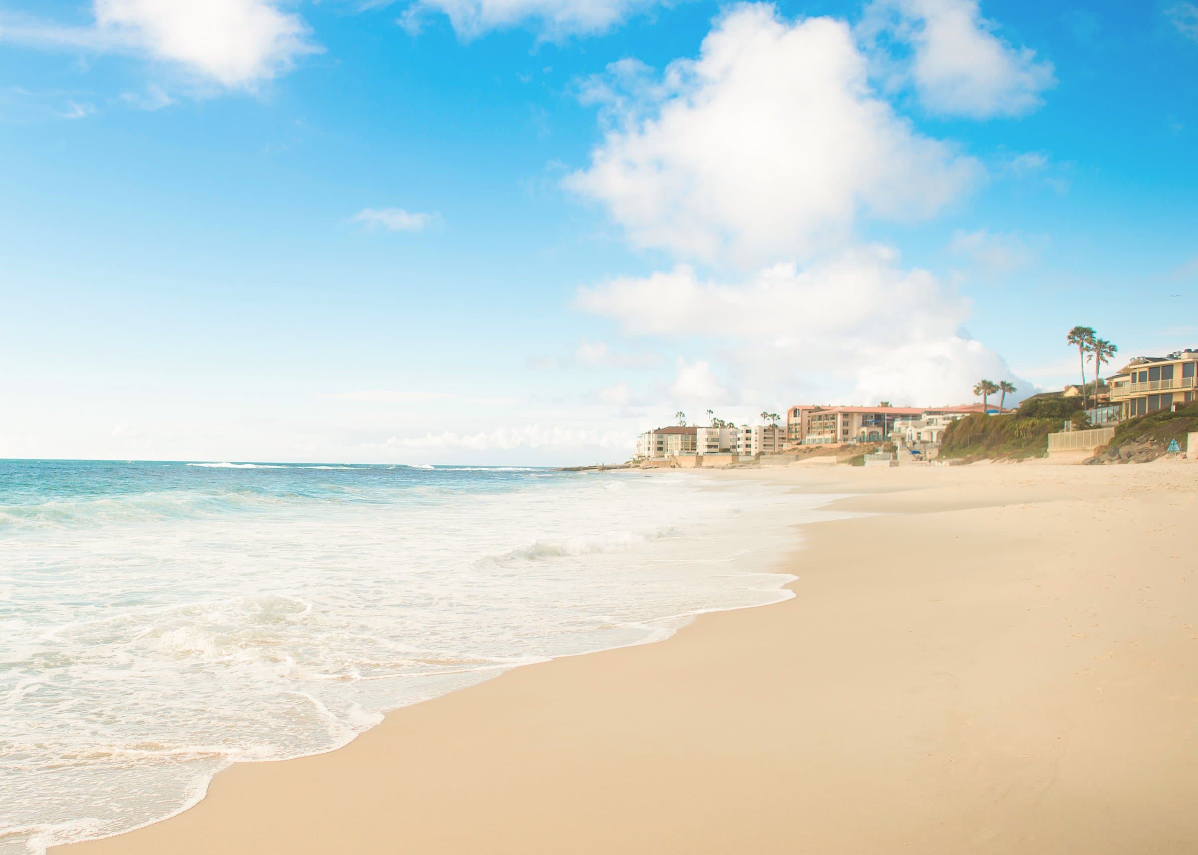 Sandy beach with turquoise surf and residential buildings along the shore under a bright sky
